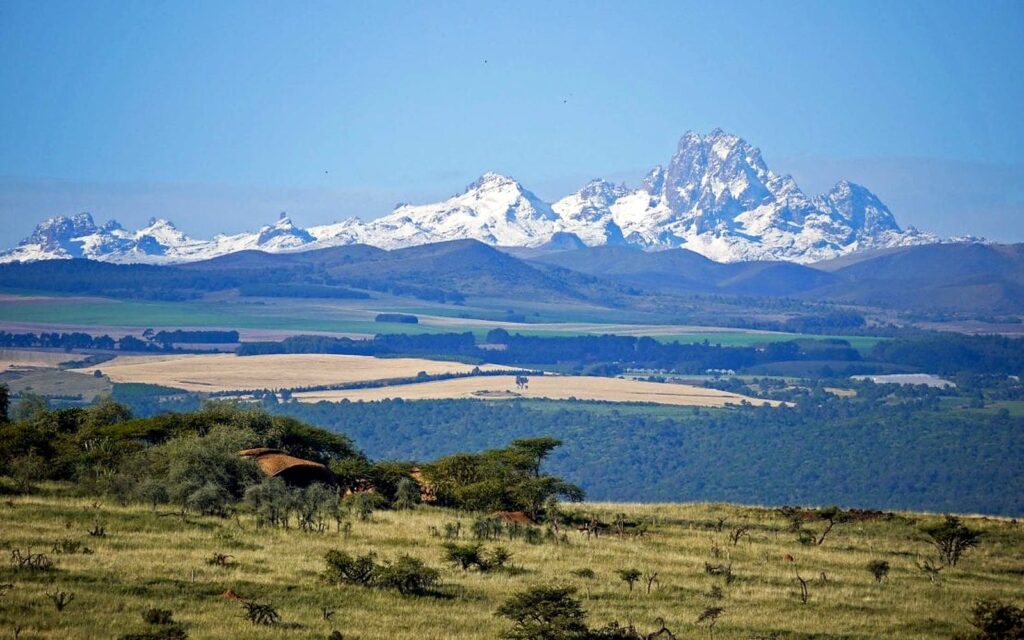 Snow-capped peaks of Mount Kenya above farmland and forest, seen from a grassy hillside